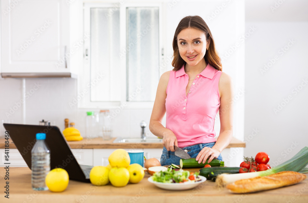 Positive woman cooking dinner and using laptop