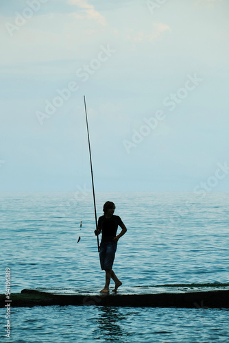 Silhouette of teenager with fishing rod on the background of the water.