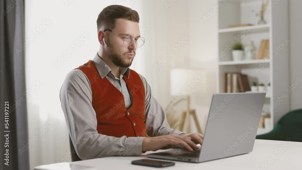 Young handsome man in eyeglasses using laptop, typing on keyboard, chatting with colleagues and listening to music, successful freelancer working online on computer Young handsome man in eyeglasses using laptop, typing on keyboard, chatting with colleagues and listening to music, successful freelancer working online on computer