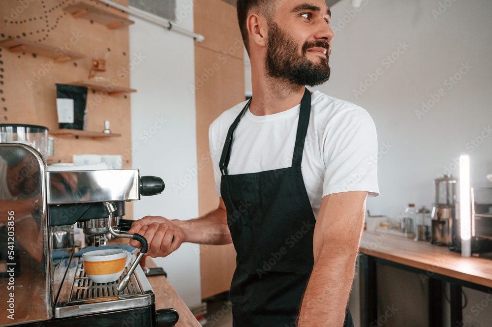 Using coffee machine. Cafe worker in white shirt and black apron is indoors Using coffee machine. Cafe worker in white shirt and black apron is indoors