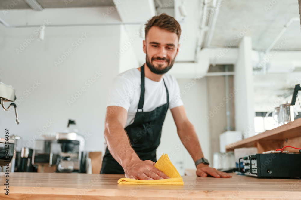Cleaning table. Cafe worker in white shirt and black apron is indoors Cleaning table. Cafe worker in white shirt and black apron is indoors