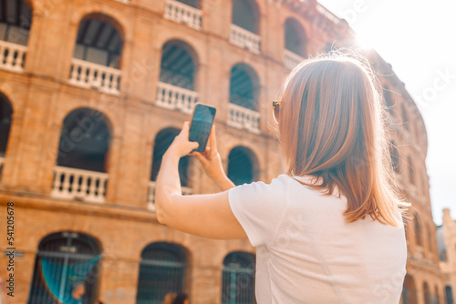 Rear view of blonde woman tourist taking photo on her mobile phone while walking on the old town of Plaza de Toros de Valencia. View from the back.  © Shi 