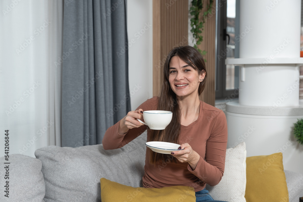 Young satisfied smiling woman drinking coffee or tea in her cozy new apartment. Female starting new life at new home sitting on sofa looking at camera. Young satisfied smiling woman drinking coffee or tea in her cozy new apartment. Female starting new life at new home sitting on sofa looking at camera.