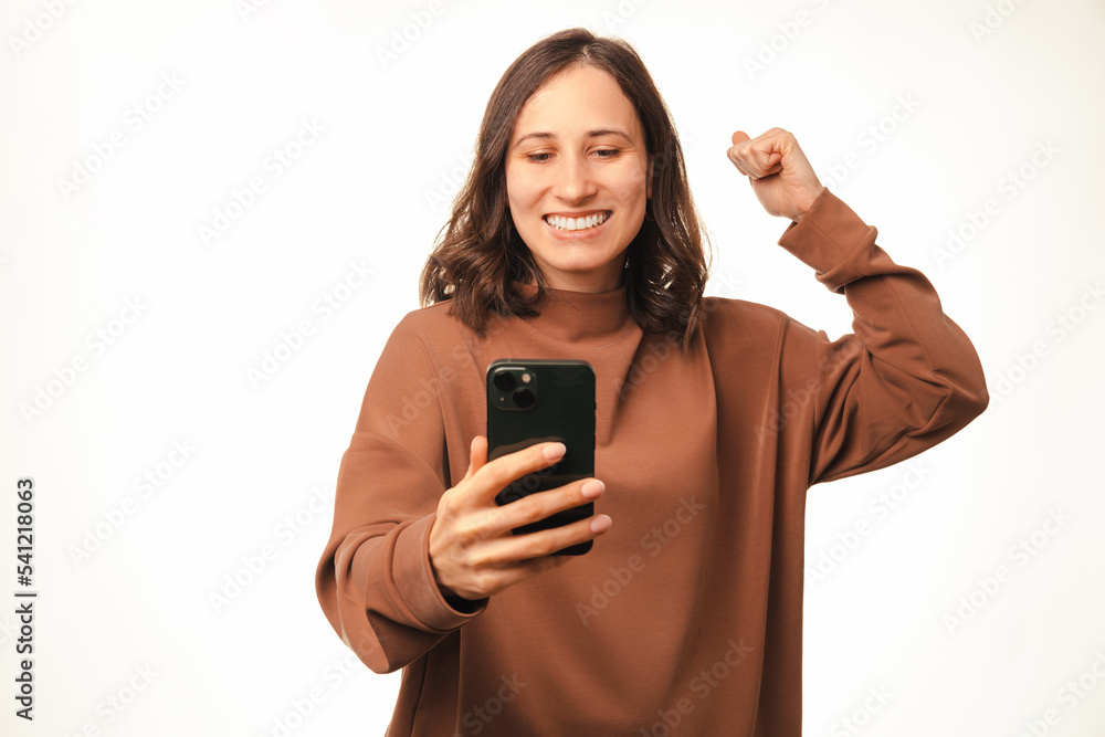 Fototapeta premium Photo of joyful young woman smiling and looking at smartphone and celebrating victory