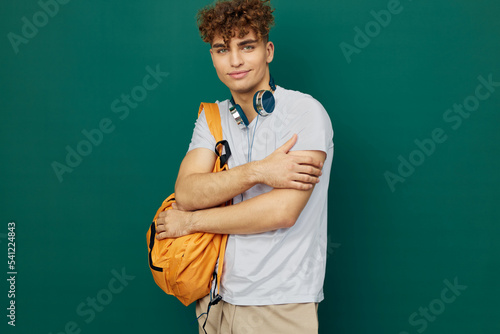a close horizontal portrait of a handsome man with curly hair standing on a green background holding headphones around his neck and clutching a bright backpack