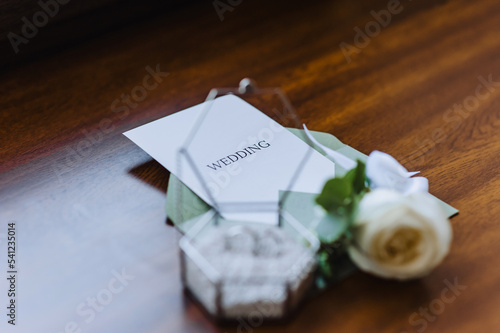 Wedding details and accessories close-up: a boutonniere, gold rings in a glass box, an envelope with a seal with a heart, a sheet of paper with an inscription. Photo, top view.