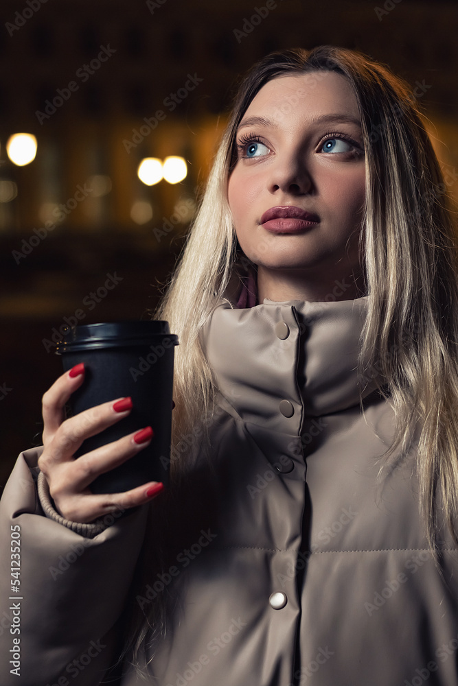 The girl walks in the night city with a glass of coffee.