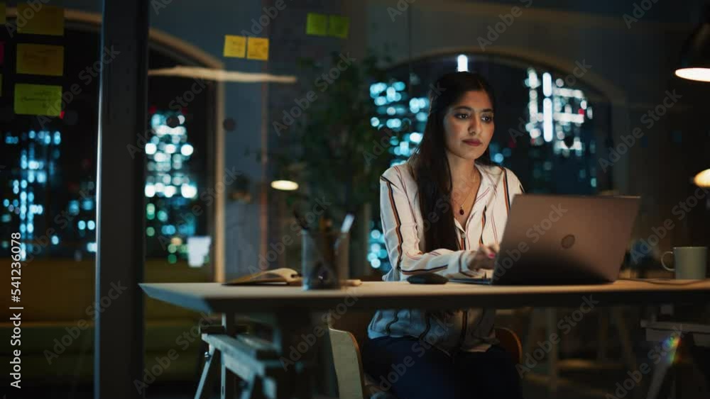 Beautiful Female in a Striped White Shirt Sitting at a Desk in Creative Office, Working on Tasks on Laptop Computer. Young Creative Specialist Writing Corporate Project Plan for Marketing Agenda. Beautiful Female in a Striped White Shirt Sitting at a Desk in Creative Office, Working on Tasks on Laptop Computer. Young Creative Specialist Writing Corporate Project Plan for Marketing Agenda.