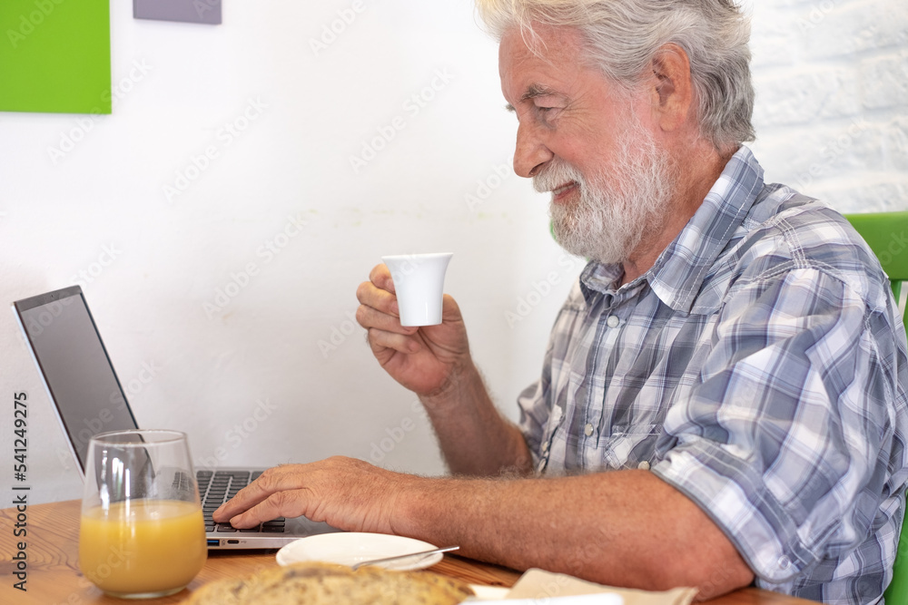Handsome senior caucasian man sitting at the table using laptop holding a coffee cup. Elderly man at breakfast time holding espresso cup while typing on computer Handsome senior caucasian man sitting at the table using laptop holding a coffee cup. Elderly man at breakfast time holding espresso cup while typing on computer