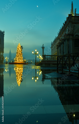 Ein Leuchtender Weihnachtsbuam spiegelt sich im Hochwasser auf dem Markusplatz in Venedig der Mond leuchtet am Himmel und im Hintergrund sieht man den Turm von San Giorgio im leuchten Morgennebel.