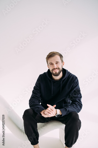 Young happy man sits and poses in armchair in studio. © Stanislav