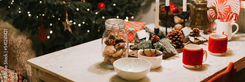 tablescapes and christmas and new year time. decorated festive kitchen cozy table with mugs  gingerbread dinner food in a jar and candles near the christmas tree. winter holiday season at home. banner