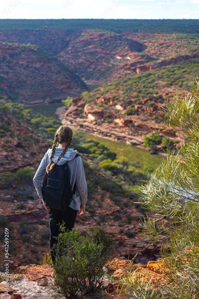 A backpacker girl stands at the top of a mountain above a river in Kalbarri National Park, Western Australia, hiking over rocks in the Australian outback