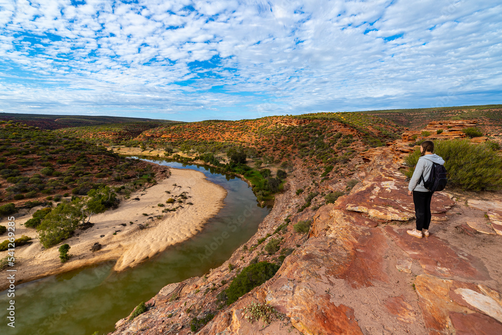 A backpacker girl stands at the top of a mountain above a river in Kalbarri National Park, Western Australia, hiking over rocks in the Australian outback