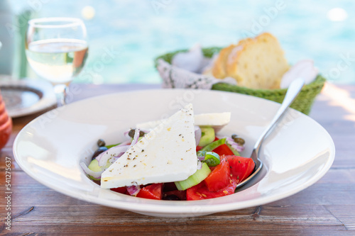 Traditional Greek Salad served in tavern, traditional greece food with Aegean sea as background. Tomatoes, cucumber, onions, olives, peppers, cappers and olive oil