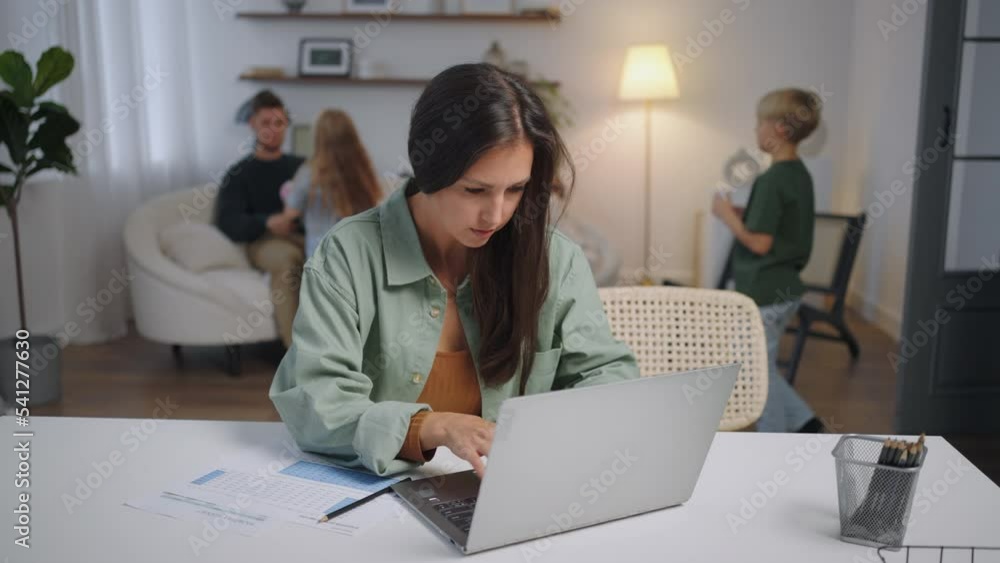 A business woman works remotely at home on a laptop while her family is singing in the background . Busy business woman. Father and children play A business woman works remotely at home on a laptop while her family is singing in the background . Busy business woman. Father and children play