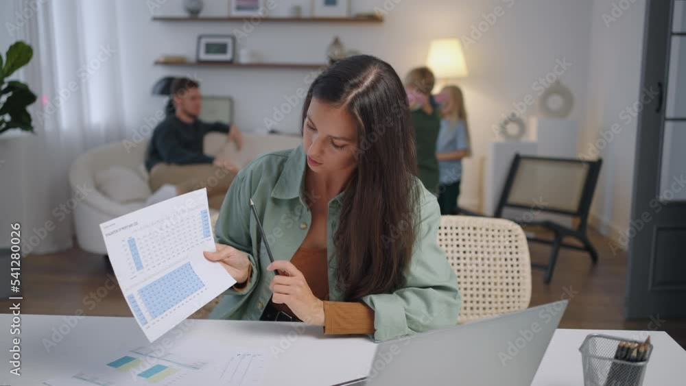 A business woman works remotely at home on a laptop and shows graphs on a webcam while the family is singing in the background . Busy business woman. Video conference A business woman works remotely at home on a laptop and shows graphs on a webcam while the family is singing in the background . Busy business woman. Video conference