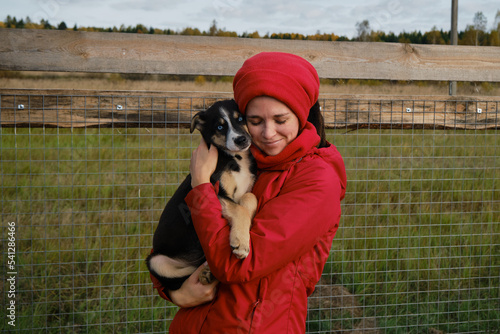 Concept of adoption of abandoned pets. Young Caucasian female volunteer at dog kennel. Woman came to animal shelter to choose puppy friend. Hold and hug blue eyed Alaskan husky puppy. photo