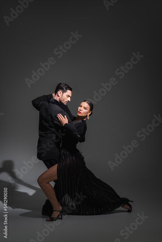 Elegant woman with red lips looking at camera while dancing tango with partner on grey background with shadow