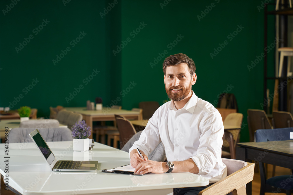 Successful businessman in white shirt working remotely with laptop, while sitting in cozy cafe. Portrait of smiling, bearded manager looking at camera, while working indoors. Concept of workspace. Successful businessman in white shirt working remotely with laptop, while sitting in cozy cafe. Portrait of smiling, bearded manager looking at camera, while working indoors. Concept of workspace.