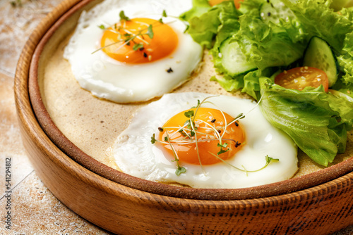 Fried eggs with micro greens, lettuce and cherry tomatoes on beige background, tile, in sunny light. Healthy breakfast is served in a wooden tray.