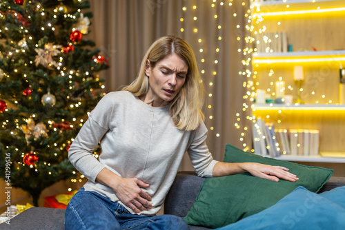 Woman abdominal pain and nausea on New Year's Eve sitting at home on the sofa against the background of a Christmas tree. Poisoning, heartburn, menstrual pain syndrome feels bad. photo