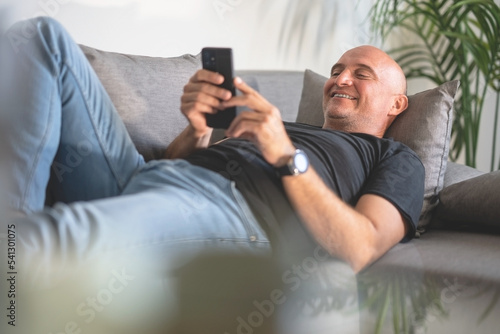 A handsome bald man is lying on the couch and reading something funny on his phone
