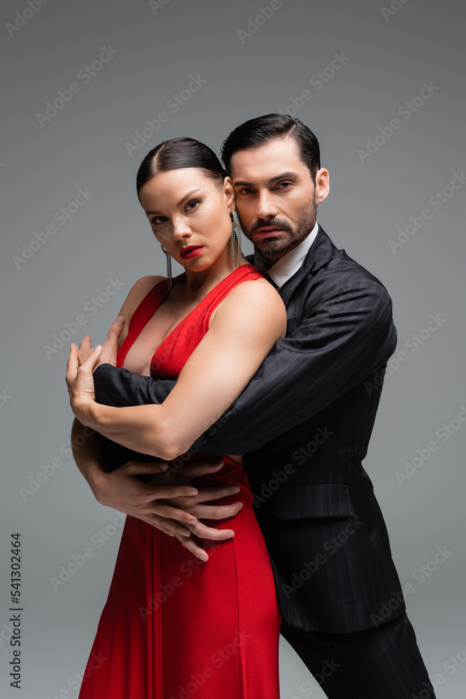 Man in suit hugging elegant woman in red dress while dancing tango isolated on grey Man in suit hugging elegant woman in red dress while dancing tango isolated on grey