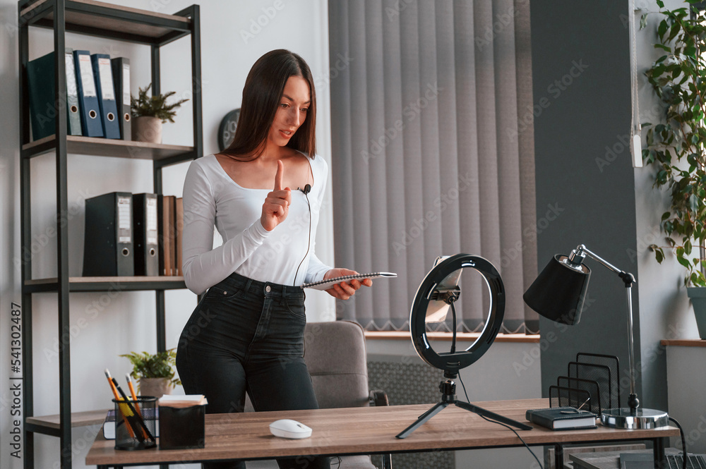 Smartphone is on the stand, recording. Young beautiful woman in white clothes is in the office