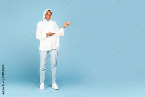 Special offer. Full length shot of black muslim lady pointing fingers aside at copy space standing over blue background