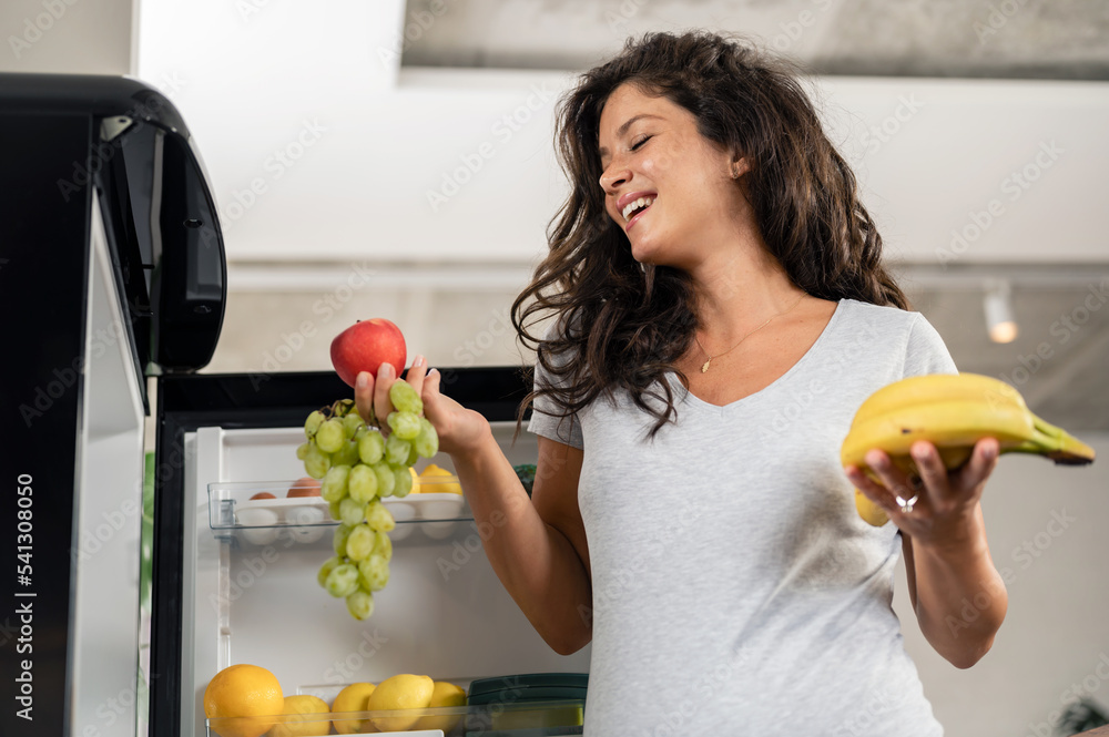 Obraz premium Beautiful young smiling pregnant woman in front of the fridge, choosing fruits to eat. Healthy lifestyle in pregnancy.