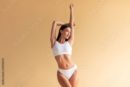 Perfect body. Slim caucasian lady posing with hands raised up, posing in white lingerie at beige background