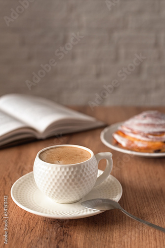 A concept for wishing a productive day. A cup of fragrant cappuccino, a cinnabon bun and a book on the wooden kitchen table. Still life in the style of minimalism. Selective focus. Space for text.