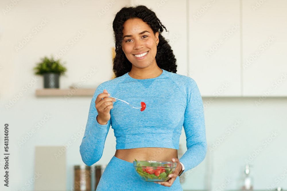 Sport and diet concept. Happy black woman eating fresh salad, standing in kitchen and smiling at camera, copy space
