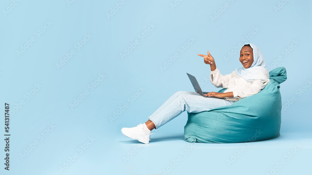 Happy black muslim woman using laptop while sitting on beanbag chair and pointing finger at free space, panorama Happy black muslim woman using laptop while sitting on beanbag chair and pointing finger at free space, panorama