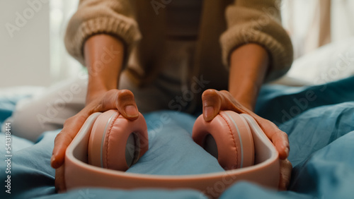 Close-up Asia girl with white cream pajamas wear pink headphone on bed happy dancing listen music smiling looking at camera in bedroom holiday morning light from window. Female morning vibes concept.