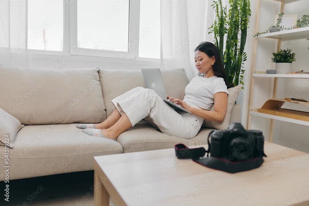 Asian woman photographer lying at home on the couch and reviewing photos from the camera in laptop for color correction retouching, work as a freelance business photographer for yourself home office Asian woman photographer lying at home on the couch and reviewing photos from the camera in laptop for color correction retouching, work as a freelance business photographer for yourself home office