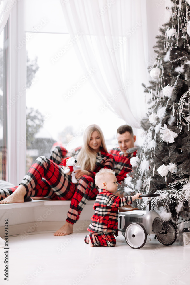 Young happy family in the same pyjamas: smiling mom, dad are sitting on big window, baby boy playing with toy retro car by the Christmas tree. The theme of the family holiday is New year and Xmas. Young happy family in the same pyjamas: smiling mom, dad are sitting on big window, baby boy playing with toy retro car by the Christmas tree. The theme of the family holiday is New year and Xmas.