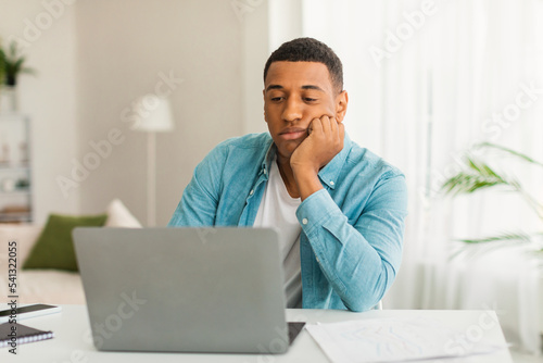 Sad tired bored young black guy in casual looks at laptop in light living room office interior, empty space