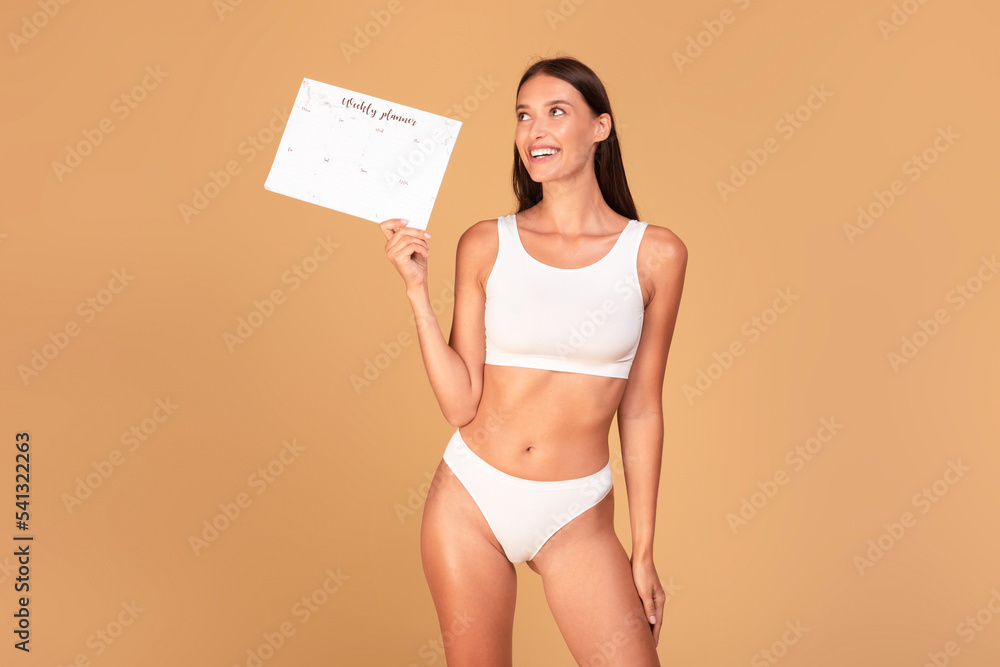 Naklejka premium Happy young woman in underwear holding calendar and smiling, posing over beige studio background, looking aside