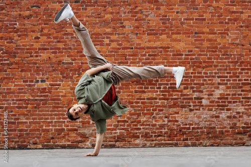 Full length shot of young man doing hip-hop handstand pose and smiling at camera against brick wall, copy space photo