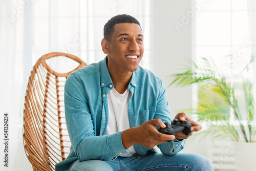 Glad happy young black male in jeans with joystick plays online game, have fun, enjoy gadget, sits in chair