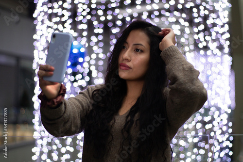Latina girl is shopping in a mall, she takes a selfie on her cell phone while touching her hair. In the background there are beautiful Christmas lights.