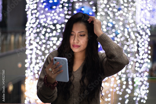 Latina girl is shopping in a mall , she smiles and looks at her cell phone while touching her hair . In the background there are beautiful Christmas lights.