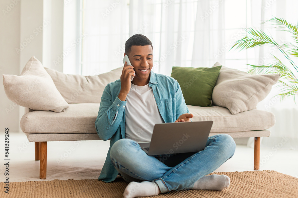Smiling busy young black male in denim calls by phone, has chat on laptop, sit on floor in living room interior Smiling busy young black male in denim calls by phone, has chat on laptop, sit on floor in living room interior