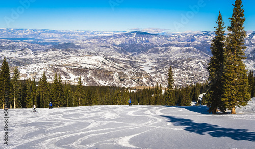 View of ski slope near Aspen, Colorado, with ski tracks on foreground and mountain range in background on sunny winter day