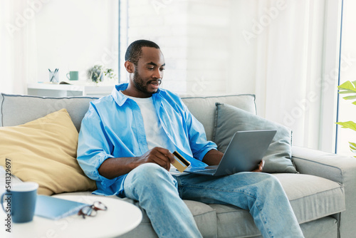 Handsome black man with laptop and credit card sitting on couch, shopping in web store from home, copy space