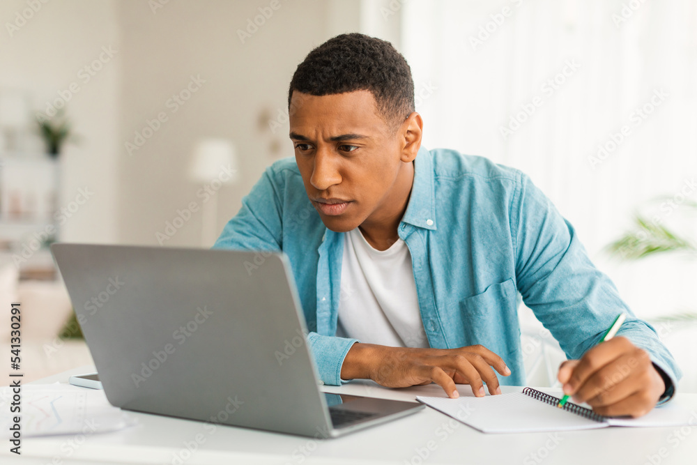 Serious concentrated bored young african american male in jeans looking at laptop making notes, find mistake Serious concentrated bored young african american male in jeans looking at laptop making notes, find mistake