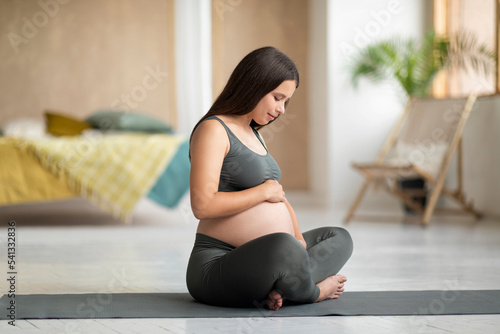 Young Beautiful Woman Expecting Baby Sitting On Yoga Mat And Embracing Belly