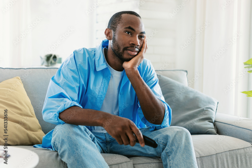 Boring television program. Discontented black guy watching TV, switching channels with remote control, sitting on sofa